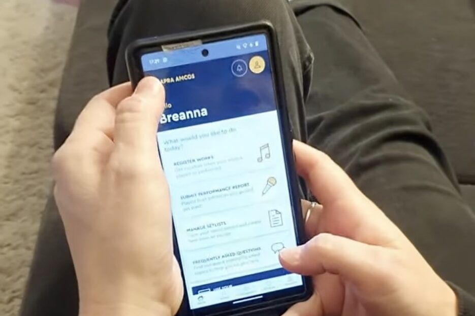 Hands holding a mobile phone. The APRA AMCOS app is open on screen.