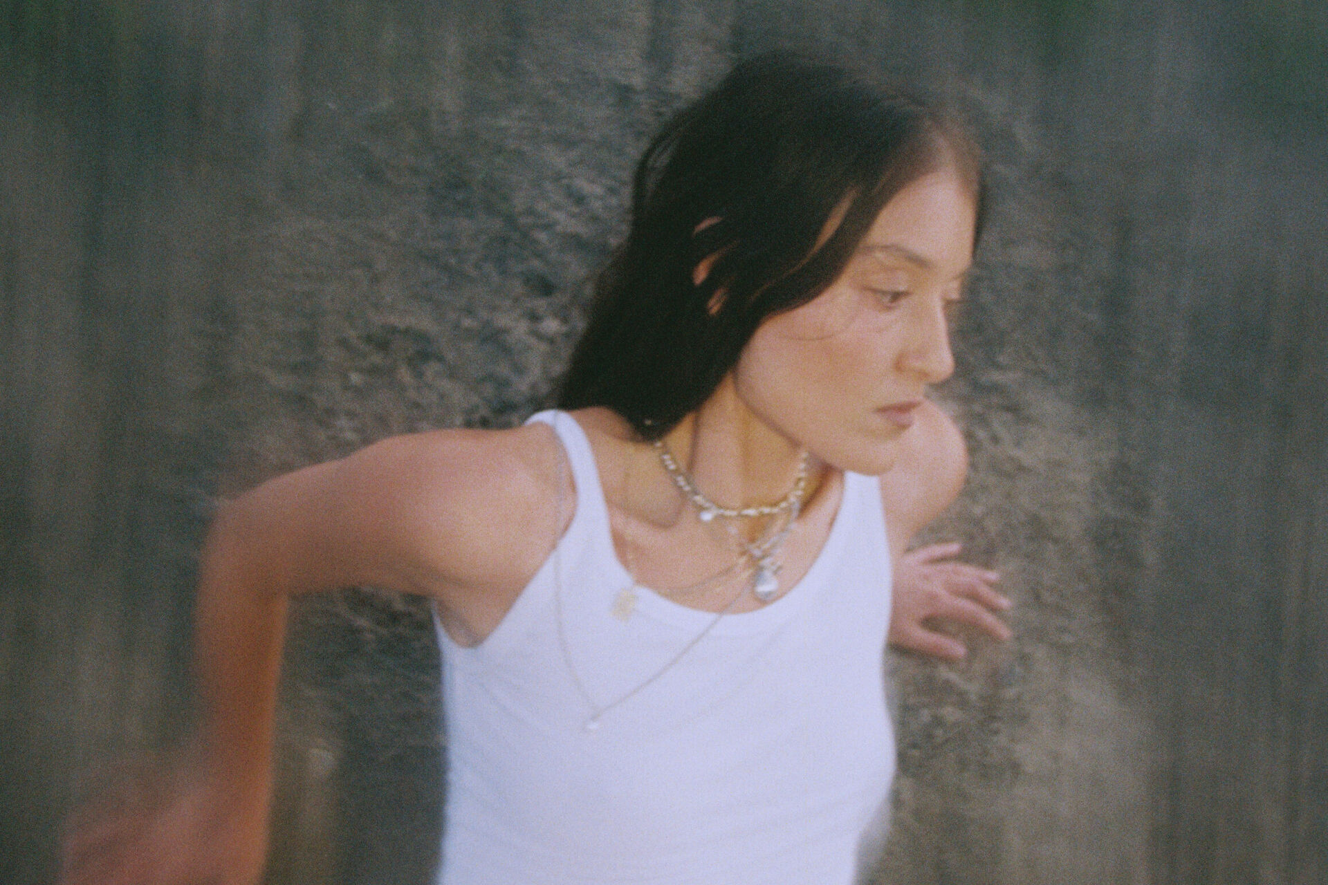 A slightly blurred style photo of Milan. She is leaning on a rock wall, her arms touching the wall. She is wearing a white singlets with layered chain necklaces. She has long dark hair and is looking away from the camera.