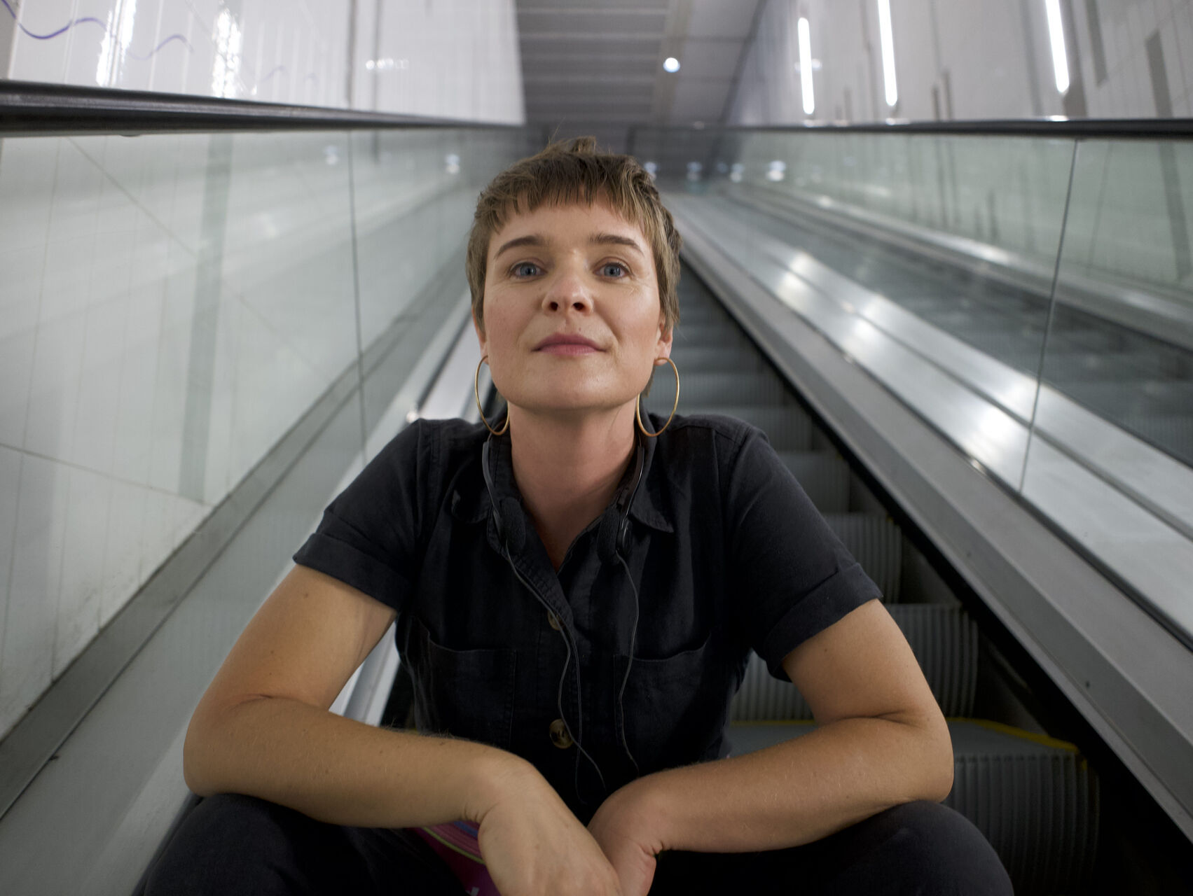 Sam is sitting at the bottom of the escalator with her arms resting on her knees. She is wearing all black and looking at the camera.