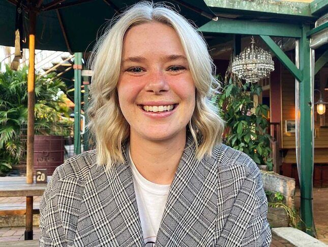 Bridie is sitting in an outdoor restaurant/pub setting. She is wearing a grey plaid blazer with a white tshirt underneath. She has blonde shoulder length hair and is looking at the camera, smiling.