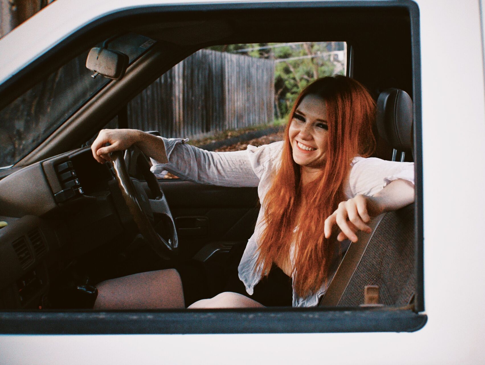 A photo of Queenie sitting in a car, taken through the passenger side window. She has one arm on the steering wheel and one resting on the back of the chair. She is wearing a white shirt and black shorts. She has long red hair and is looking at the camera, smiling.