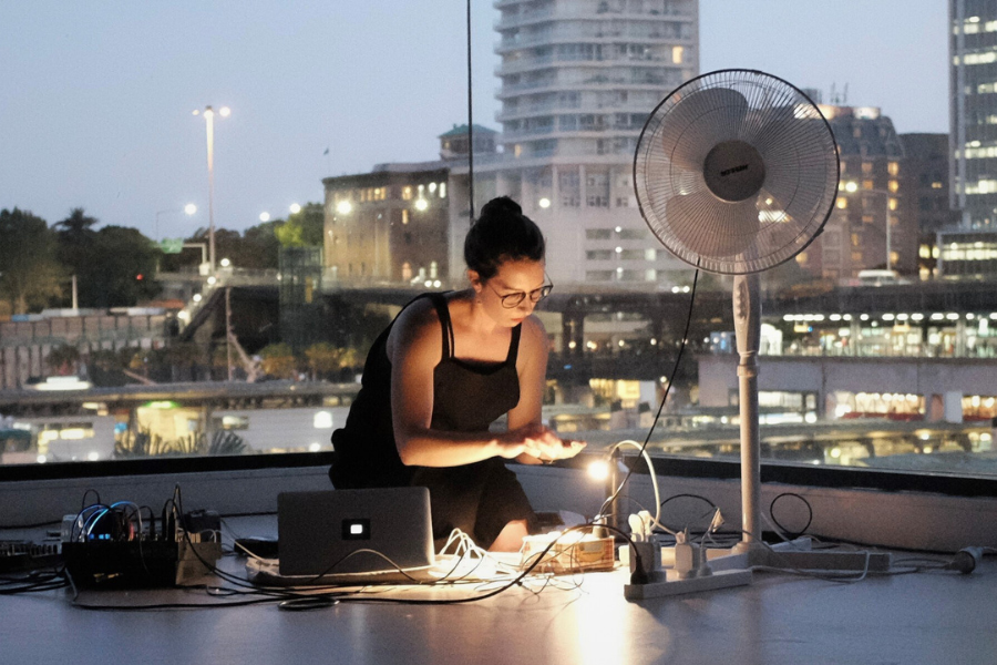 Photo of Alexandra spence performing at the MCA in Sydney. She is kneeling down, with a computer and cables. There is a small desk light shining, and a fan. In the background there are Sydney's Circular Quay skyscrapers.