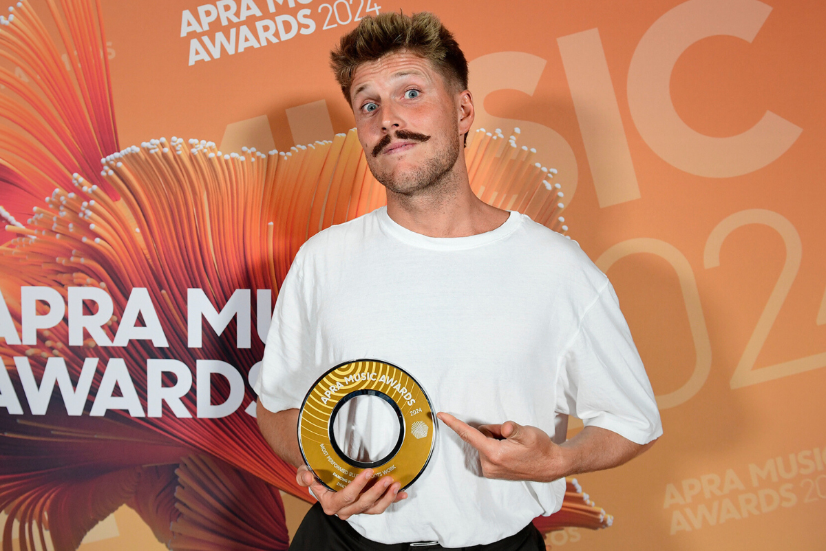 Photo of Ziggy Alberts holding an APRA Music Award, standing in front of an orange APRA AMCOS media wall. He has short blonde hair, a moustache, and is wearing a white t-shirt.