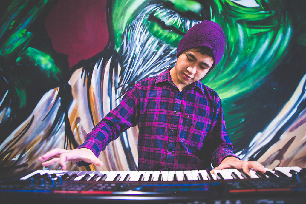 Julian Sanchez is standing playing the keyboard, wearing a purple and blue flannel button up shirt and a purple beanie. There is a painted mural on the wall behind them.
