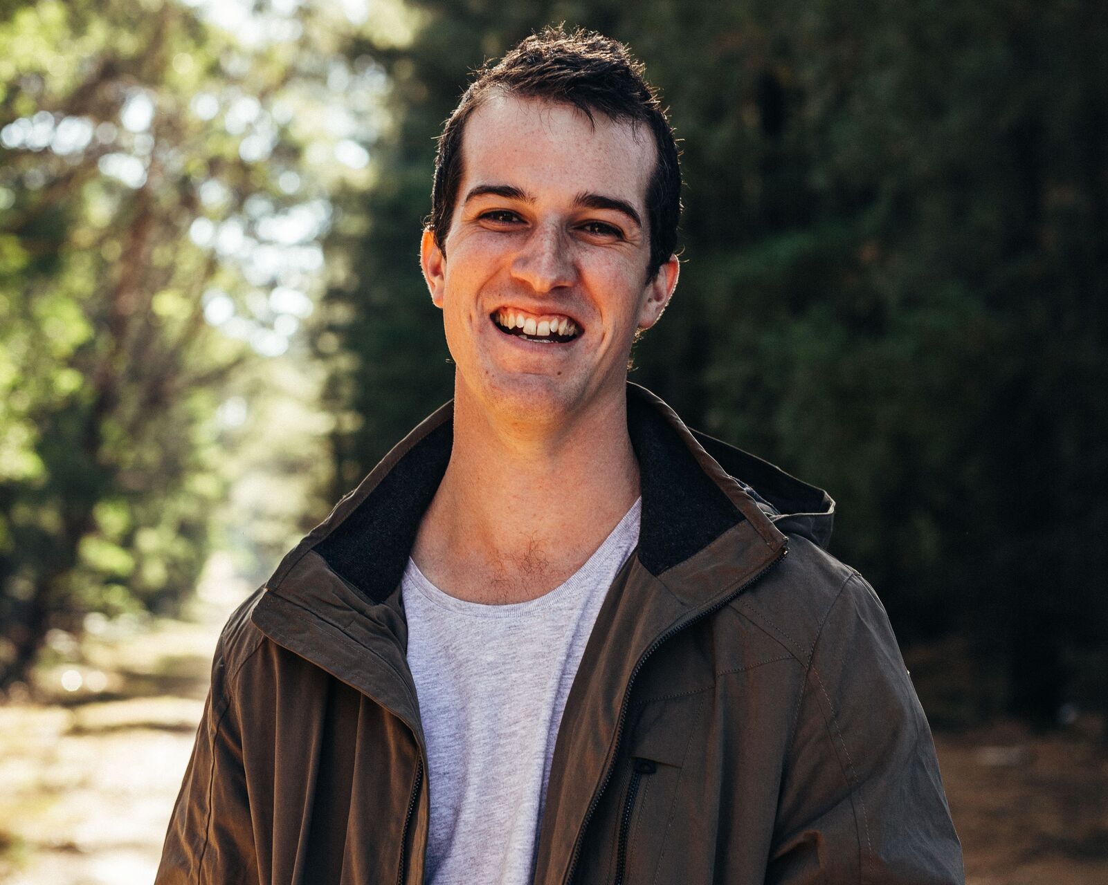 Scott stands with trees in the background. He's smiling and wearing a light grey tshirt and a dark green jacket. Scott has short dark brown hair.