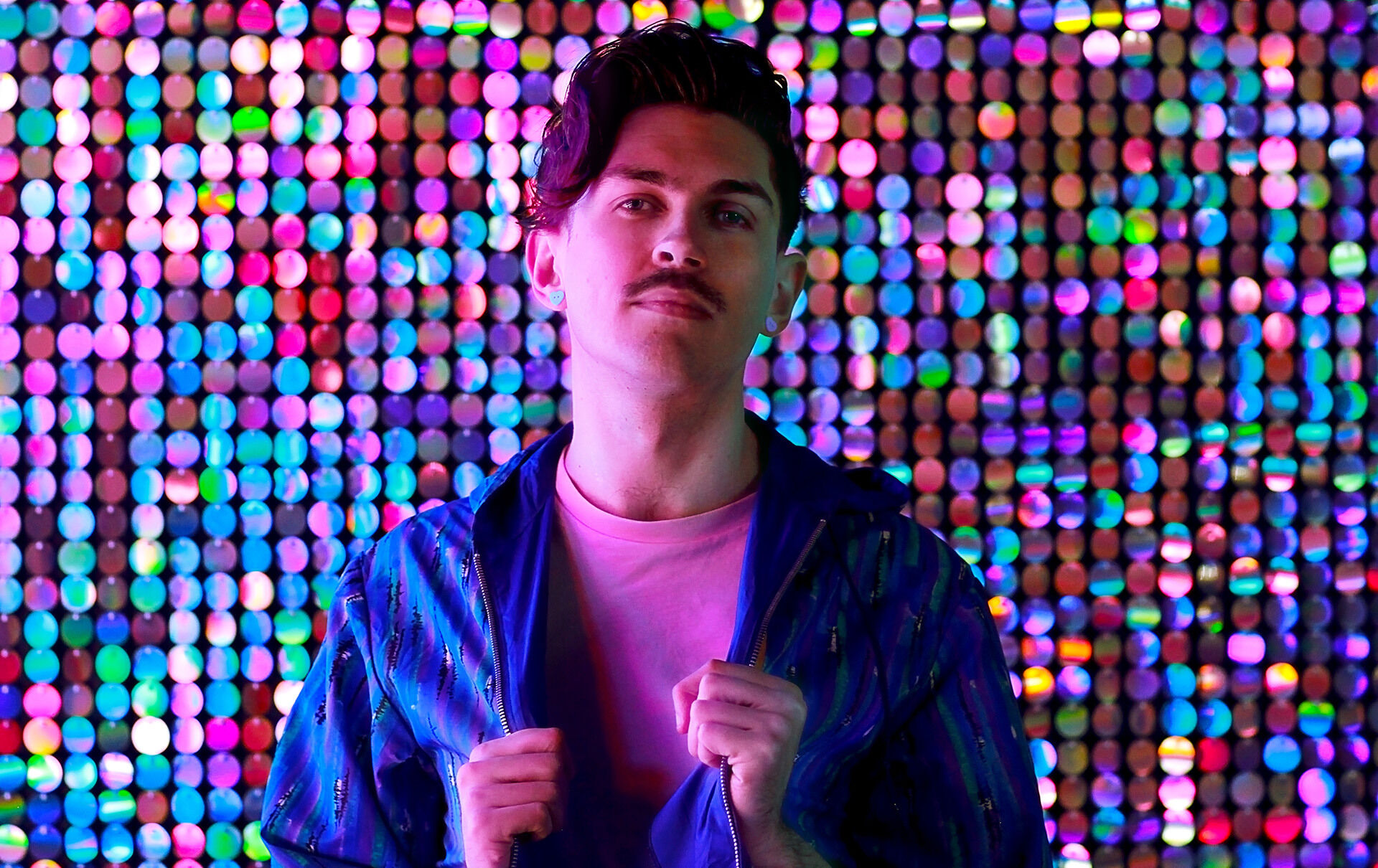 Jacob is standing in front of a colourful glittery backdrop with a blue jacket and a pink tshirt underneath. He is looking at the camera and his hands are clutching both sides of the jacket.