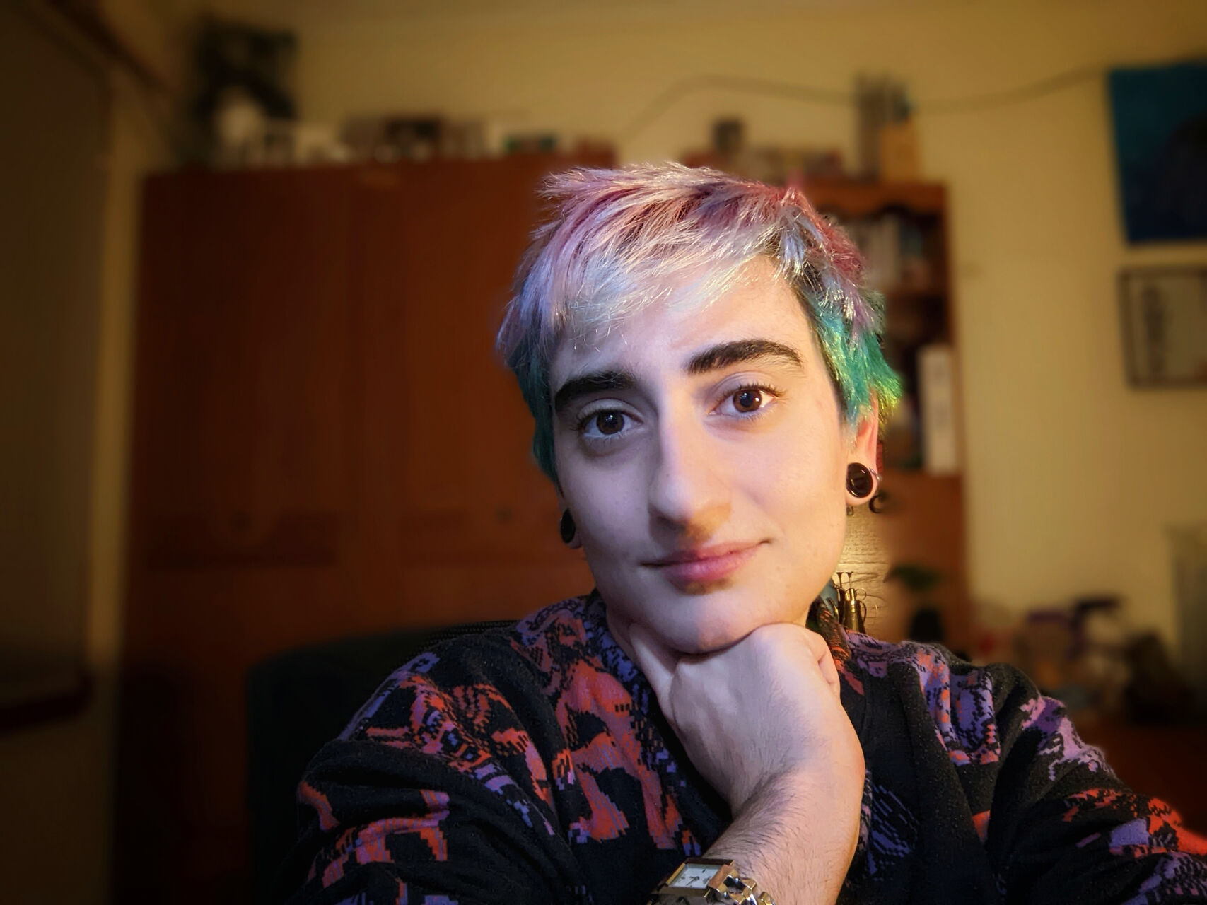 Rebecca is sitting in a room with a brown wardrobe in the background. They have short blonde hair with pastel pink, blue and green sections throughout. They're wearing a black jumper with pink and purple patterns, black circle earrings and are resting their chin on their hand.