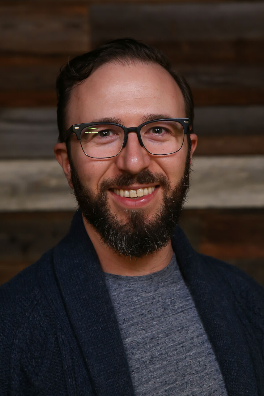 Darren is smiling, looking at the camera. he has a dark brown beard and moustache. Darren is wearing a light grey t-shirt with a dark blue cardigan blazer over the top. There is a wooden panel wall in the background.