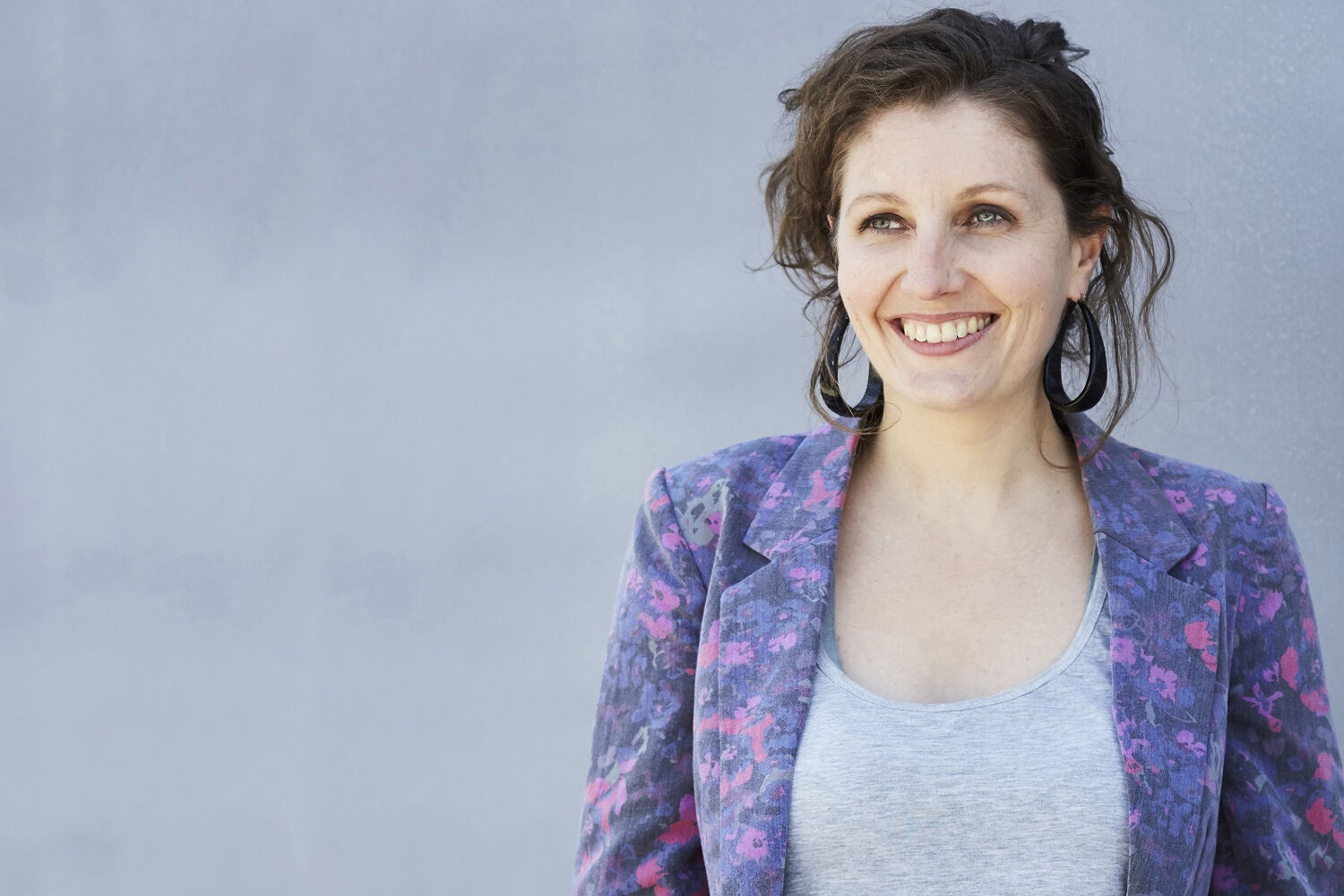 Belinda is standing in front of a grey wall, looking past the camera and smiling. She has dark brown hair which is loosely tied up and is wearing large black hoop earrings. She is wearing a light grey tshirt with a purple and pink patterned blazer over the top.