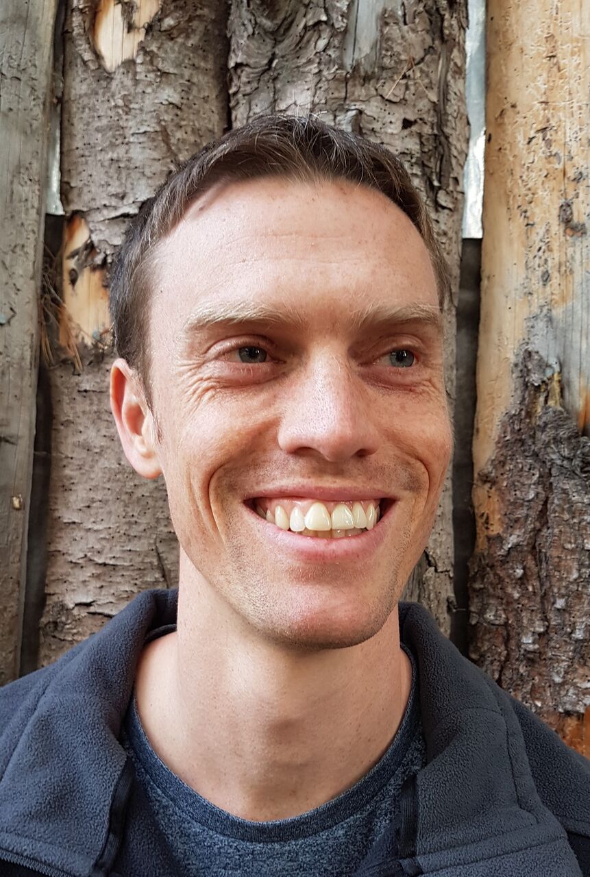 Cedar is standing in front of a tree, glancing past the camera, smiling. He's wearing a grey tshirt with a grey jacket over the top.