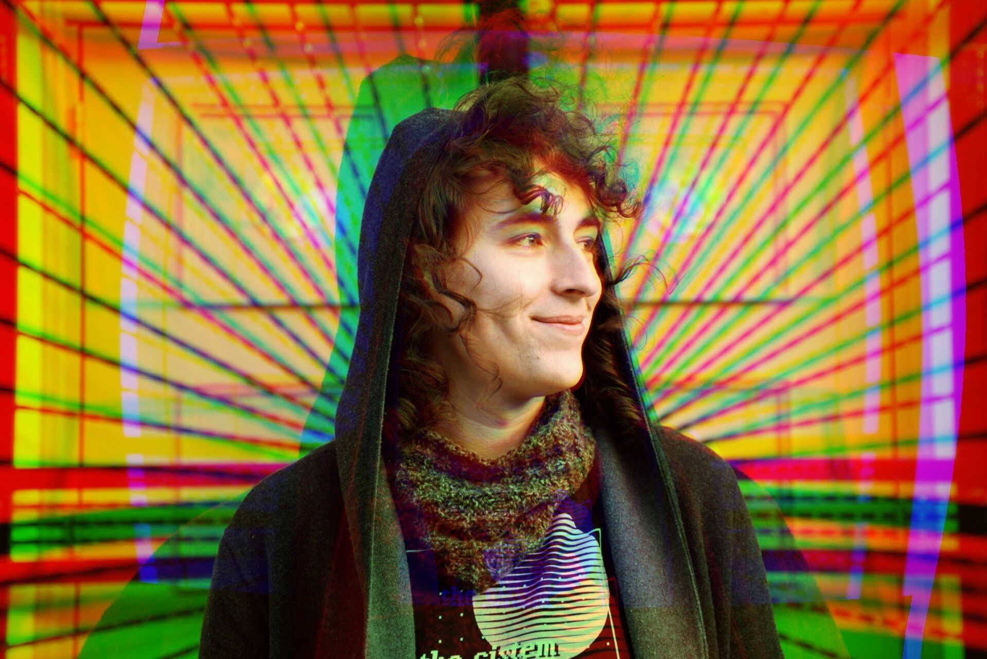 Josie is looking sideways, smiling. They are wearing a hooded open jumper with a scarf around their neck. The background has been edited to be multicoloured background.