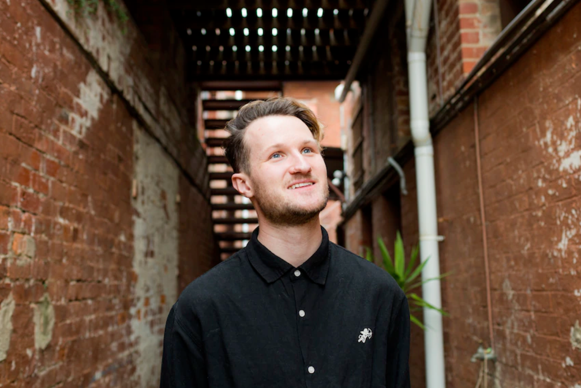 Julian is glancing upwards and is smiling. He is wearing a black button up shirt and is standing in an alleyway with brick walls either side of him.