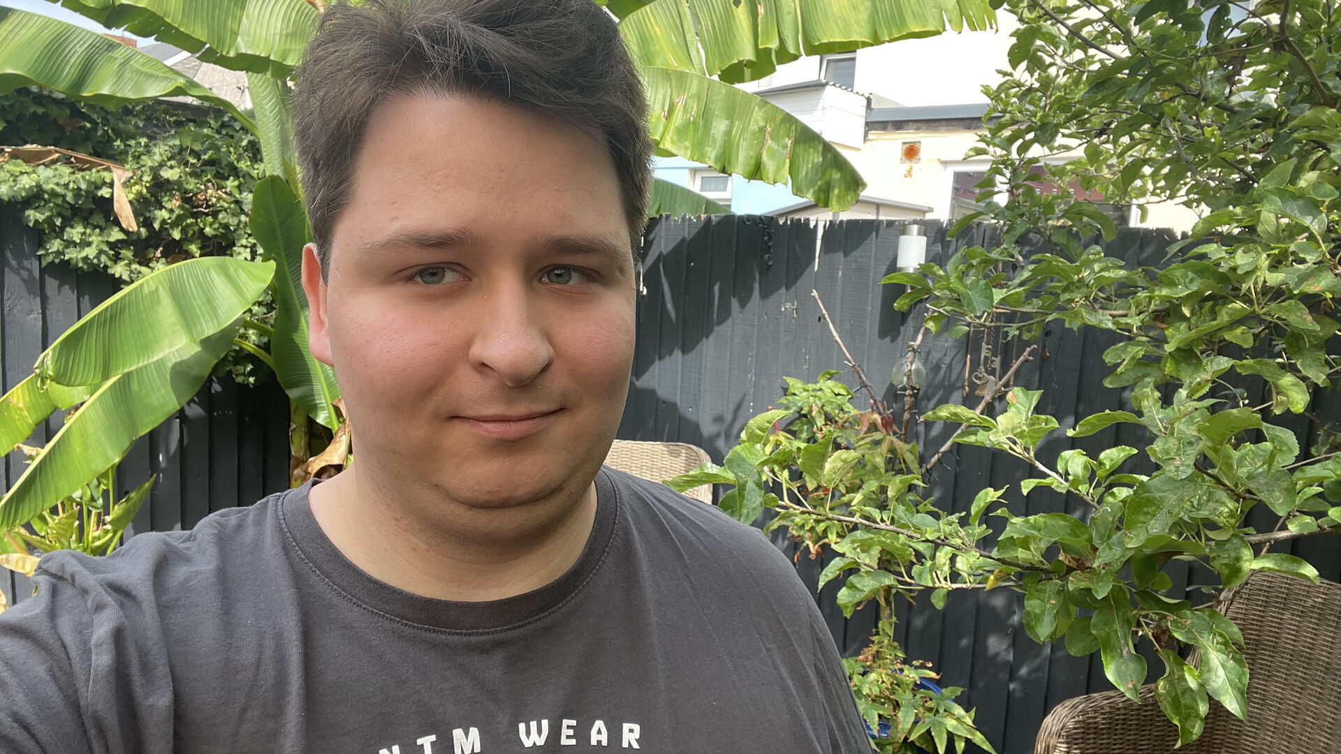 Taliesin is standing in a garden, looking at the camera. They are wearing a grey tshirt.
