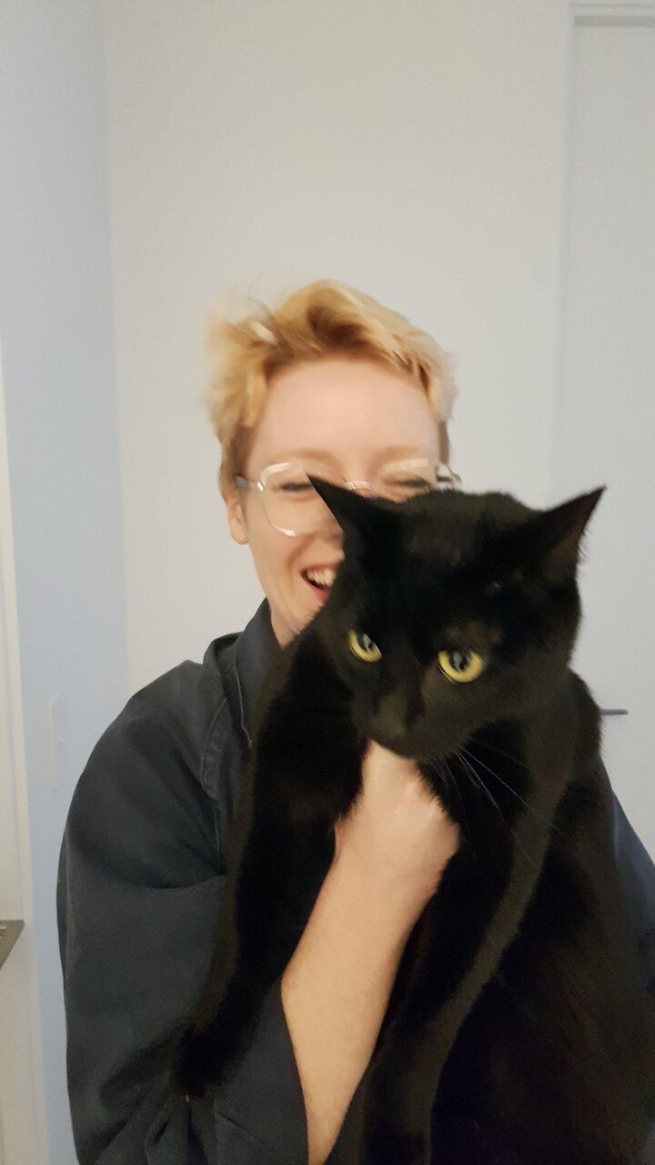 Jini has short strawberry blonde hair and is laughing while holding a black cat up to the camera.