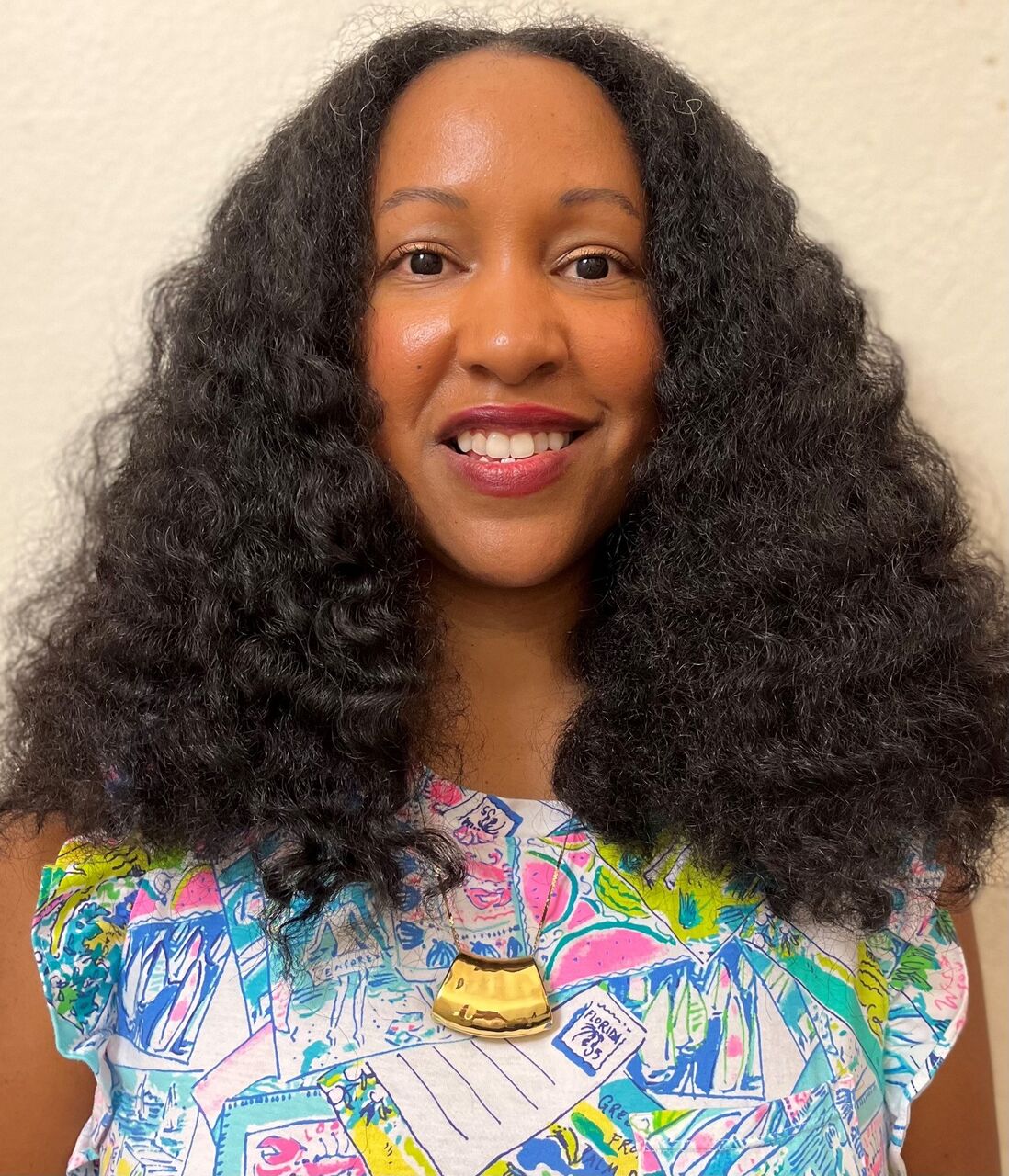 Lori is looking at the camera, smiling. Lori's hair is out and wavy. Lori is wearing a colourful patterned top and a necklace.