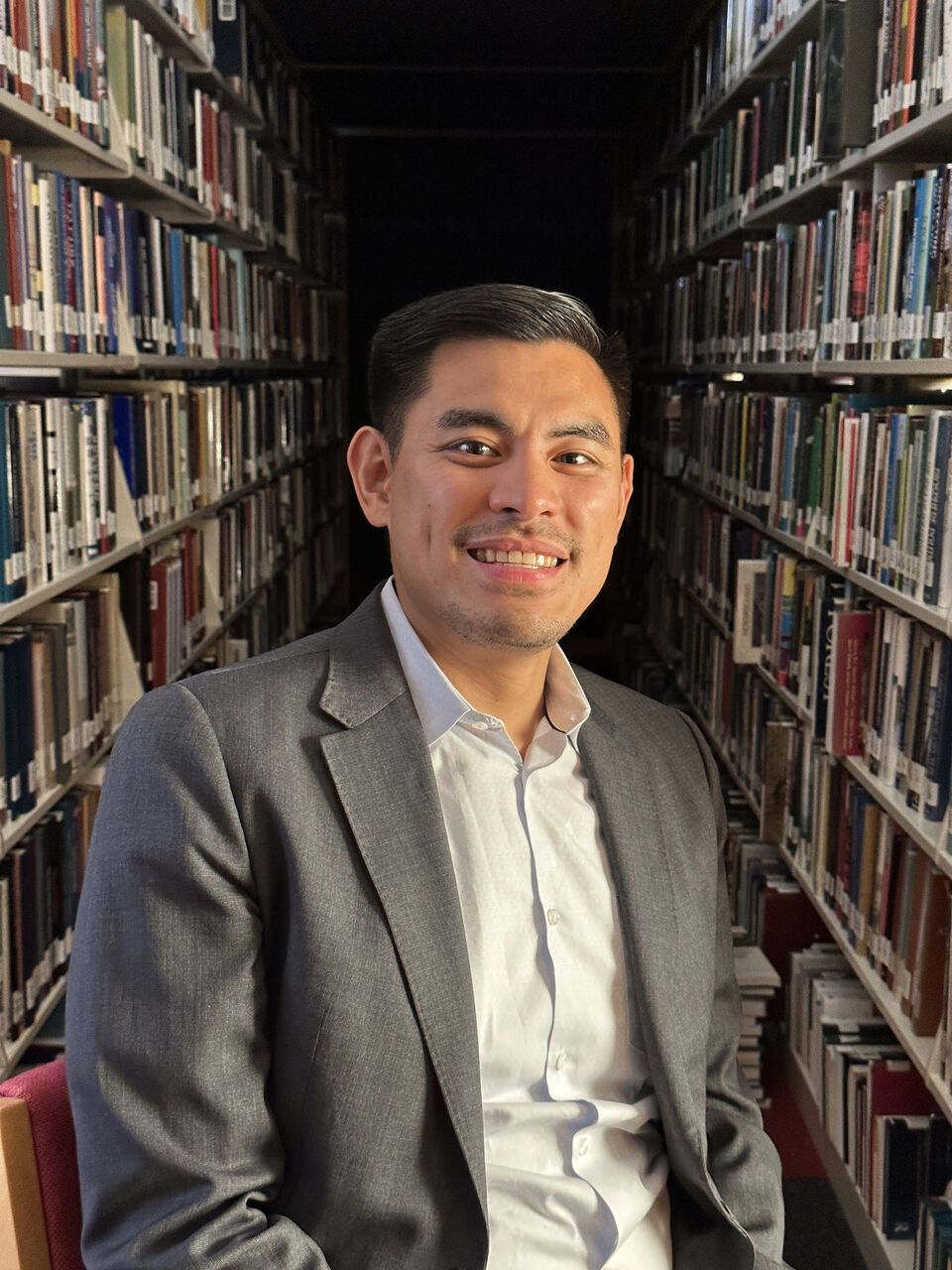 Alexander is sitting in a library. He is looking at the camera smiling. He has short black hair and is wearing a grey blazer over a white shirt.