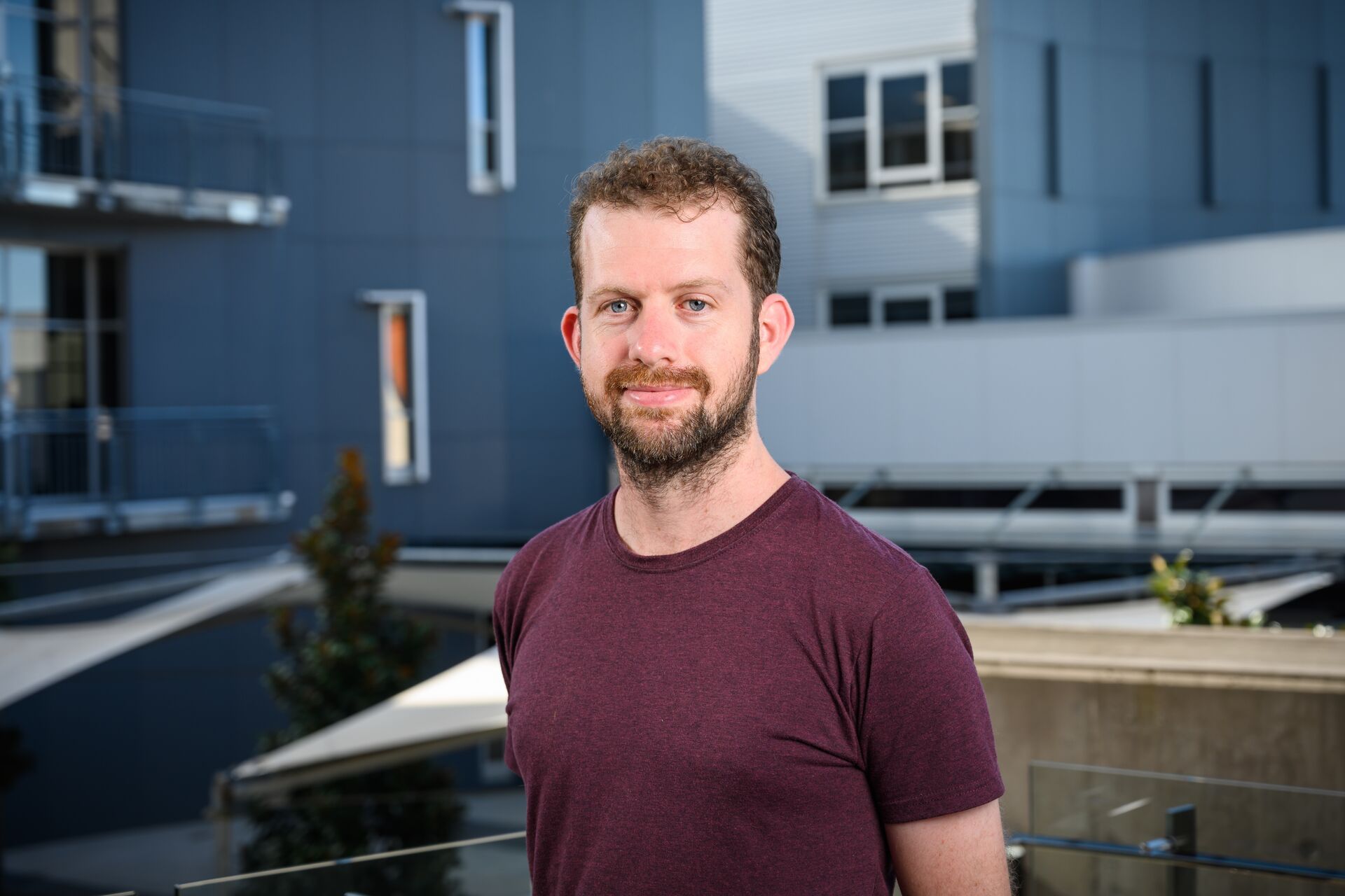 Brendan is standing in front of a building landscape. He is looking at the camera, smiling and wearing a maroon tshirt.