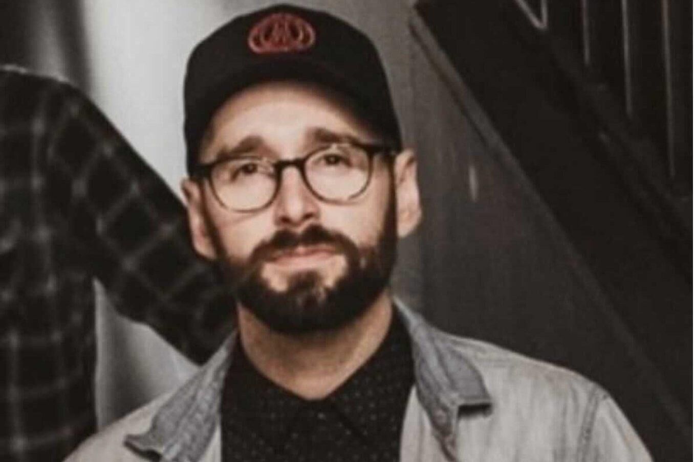 Chris is looking directly at the camera. He is wearing a black cap, a black tshirt with an open grey shirt over top and has a beard