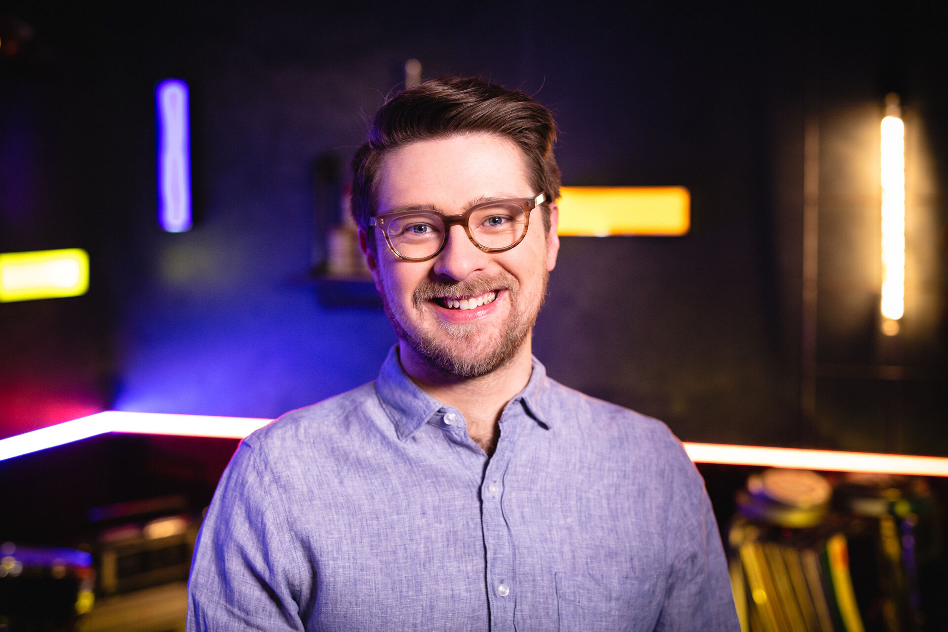 Dan is standing in a studio looking at the camera. He has short brown hair, is wearing glasses and a blue shirt.