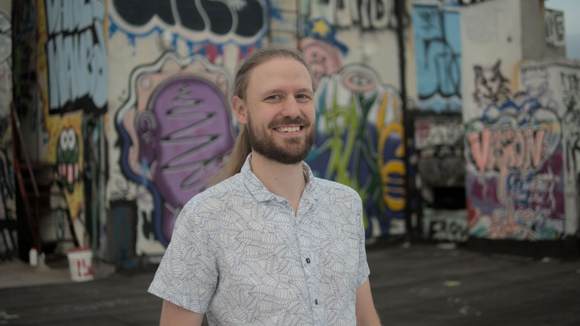 Jacques is standing in front of a graffitied wall, looking at the camera smiling. He has long hair pulled back and is wearing a light coloured shirt.