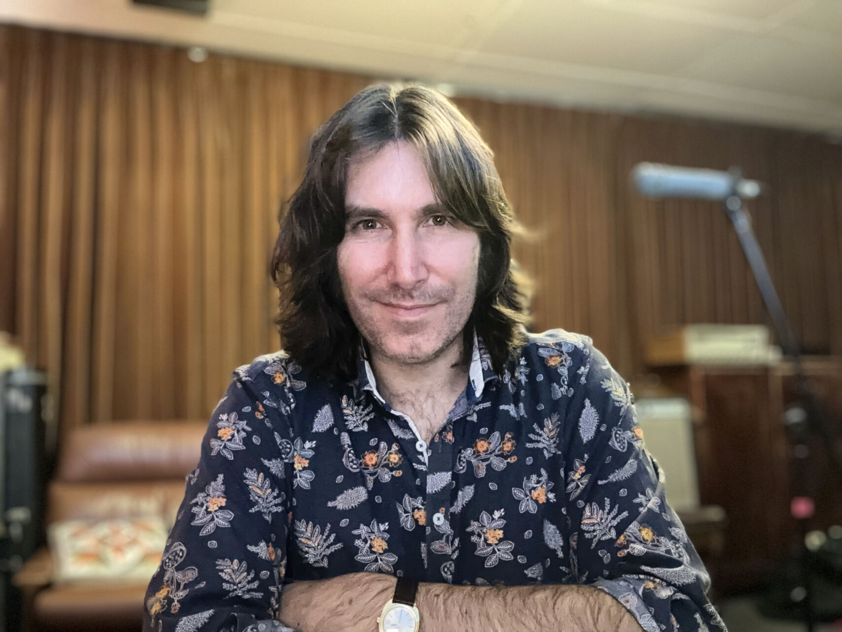 John is in a studio. He is looking at the camera with his arms crossed. He has long dark hair and is wearing a blue patterned shirt.