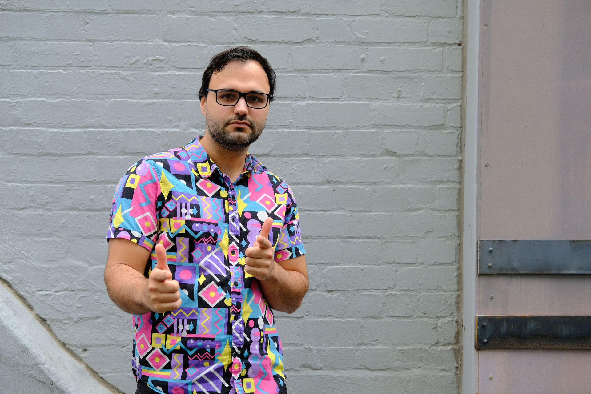 Mitchell is standing against a grey painted brick wall in a 90s geometric style shirt of pink, blue, purple, yellow and back. He is doing finger pointers at the camera and is wearing glasses.