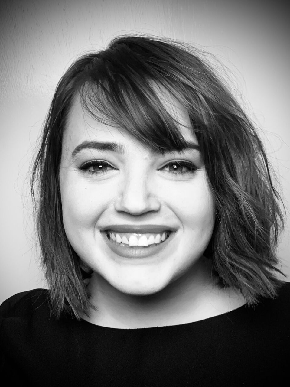 Black & white photo of Natalie. She is looking at the camera & smiling. She has dark hair with a bob and is wearing a black top.