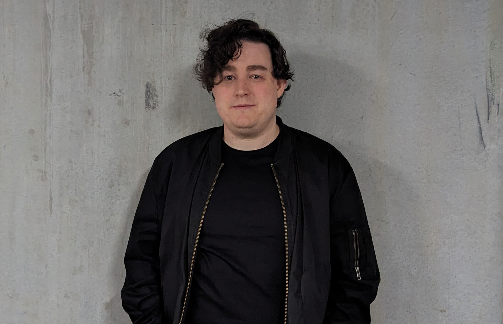 Nicholas is standing against a grey cement wall with hands in pockets. They have short dark hair and are wearing a black tshirt with a black jacket.