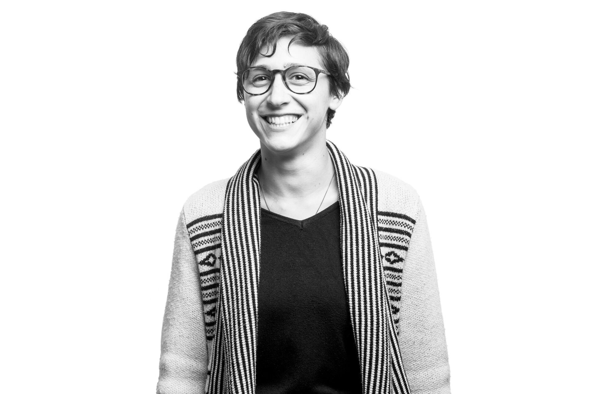 Black and white photo of Talia from torso up. They are looking at the camera and smiling. They have short dark hair and are wearing glasses.