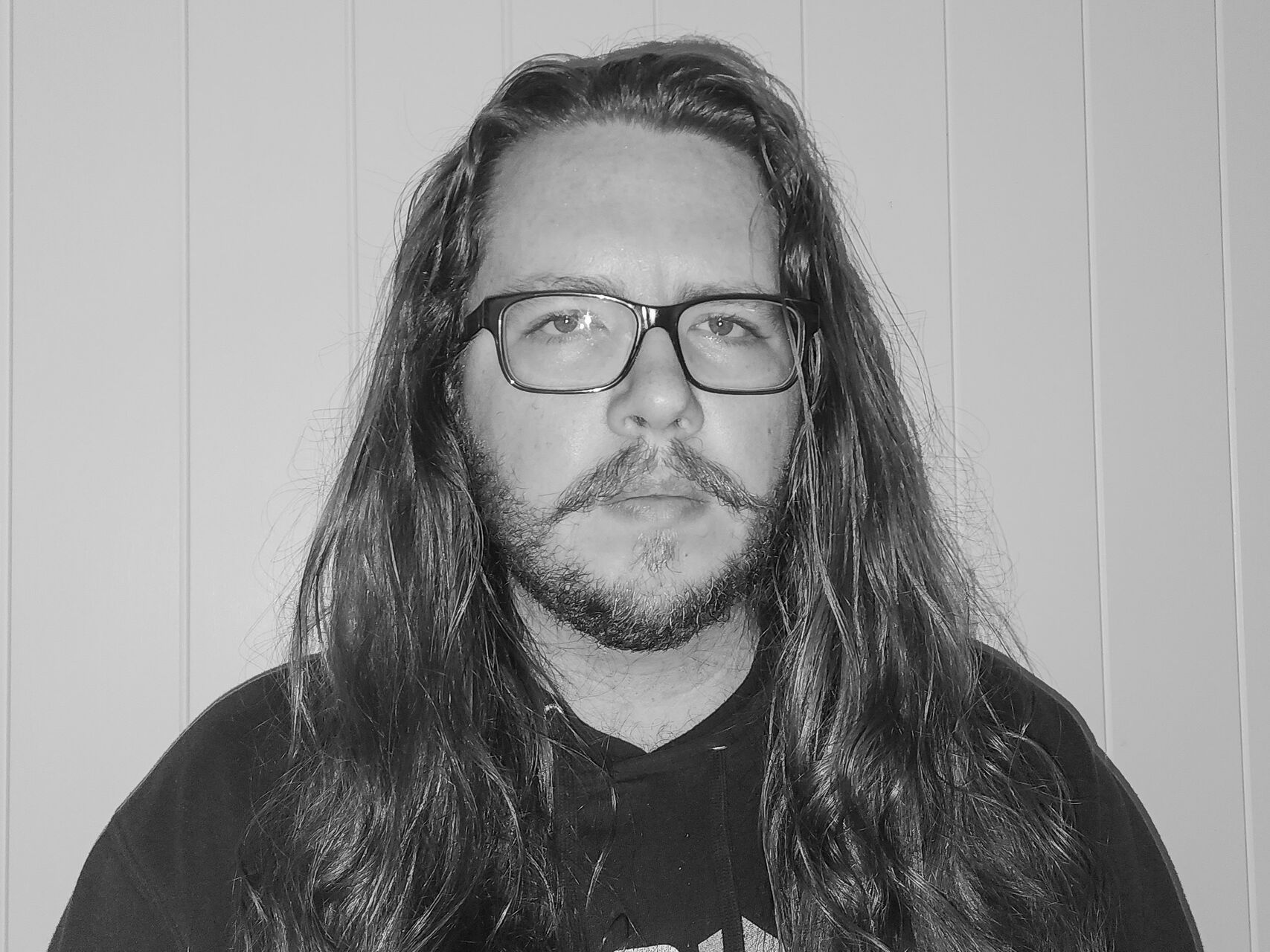 Black & white photo of Tim Sunderland. He is wearing glasses and looking at the camera. He has long dark hair with a moustache and beard. He is wearing a black tshirt.
