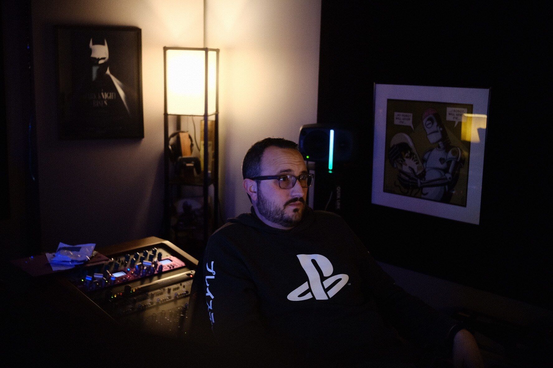 Blake is sitting in a dimly lit studio, wearing a PlayStation hoodie. He has dark hair and is wearing glasses.