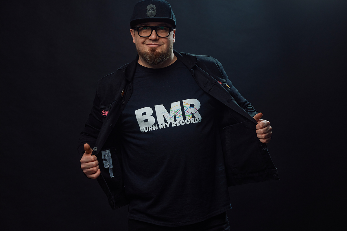 Jayde is looking at the camera in front of a black background. He is wearing a bomber style jacket and holding it open to reveal a black tshirt with BMB Burn My Record written on it in white. He is wearing a black cap with glasses.