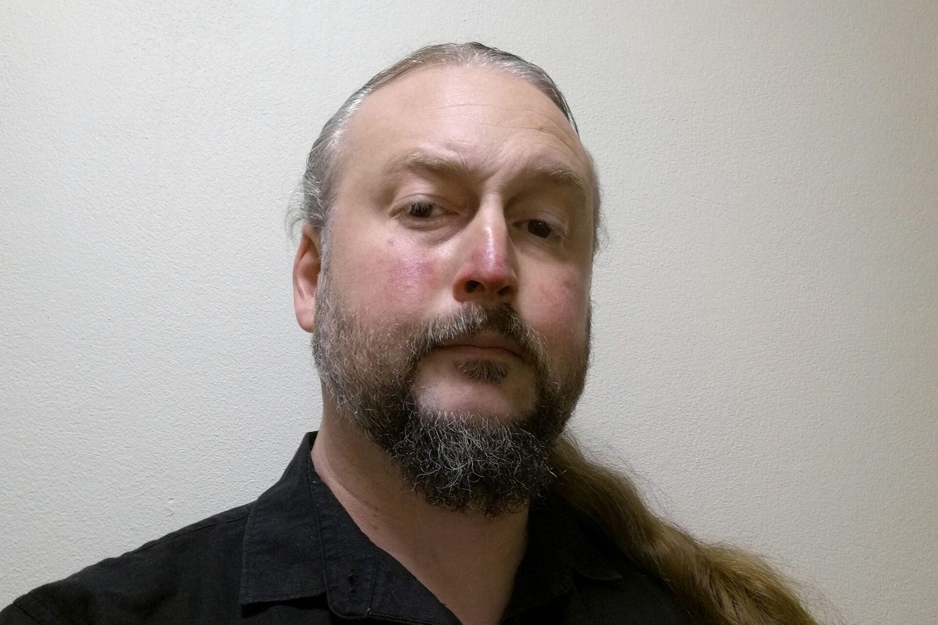 Martin is looking at the camera, wearing a black shirt and has a long blonde ponytail.