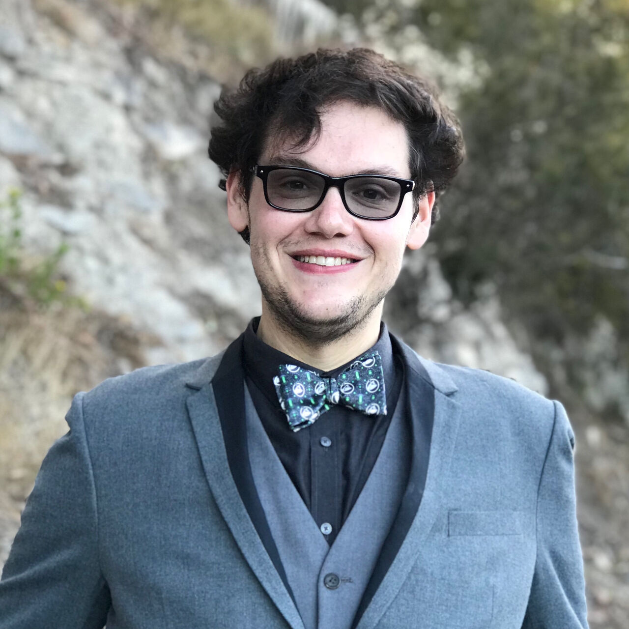 Paris is standing in front of an outside landscape and is looking at the camera, smiling. He is wearing a grey suit with black shirt and a blue patterned bow tie.