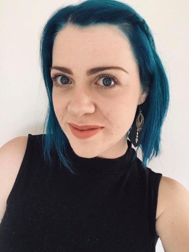 Taylor is standing against a white wall, looking at the camera. They have blue shoulder length hair and are wearing a black sleeveless top.