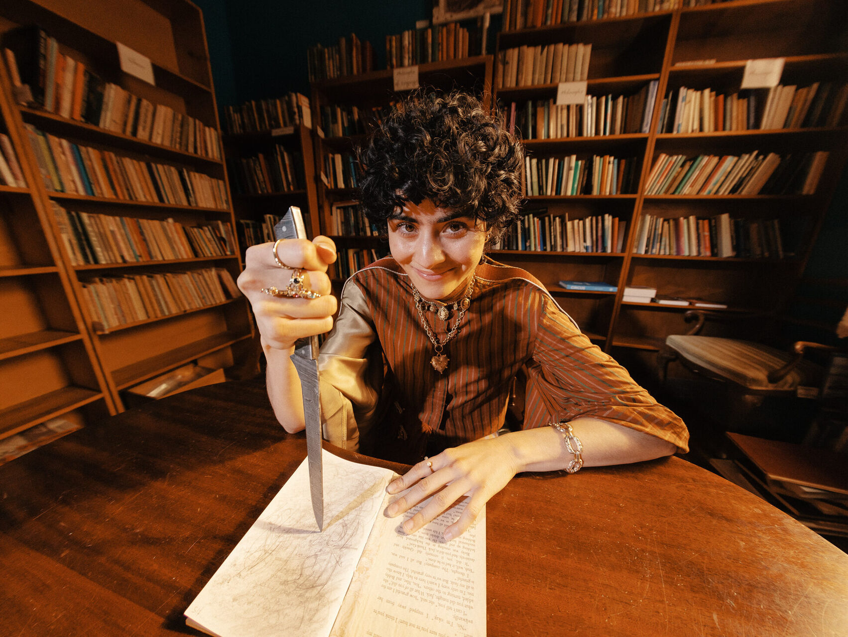 Montaigne is sitting at a desk in a library with a book, holding a knife. They have short curly brown hair and are looking at the camera.