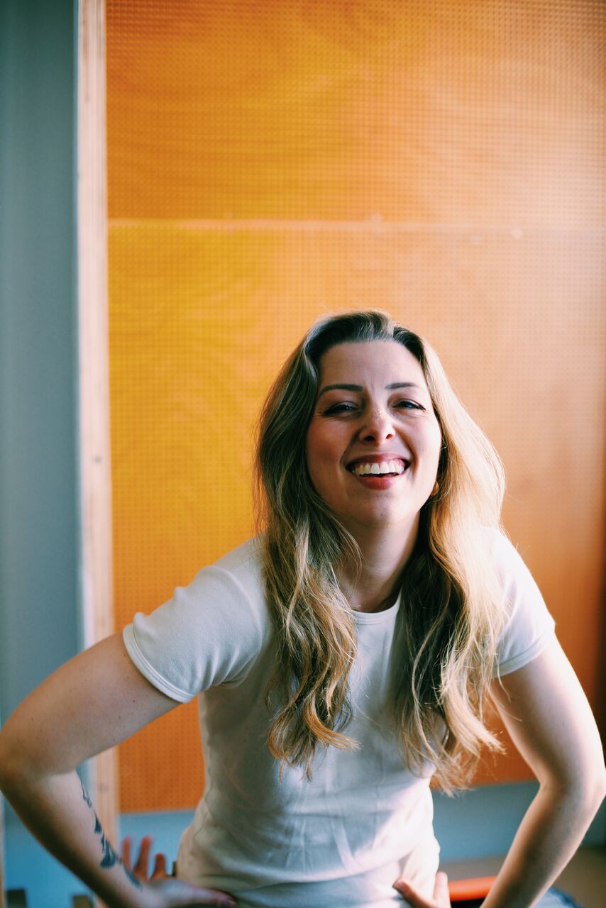 Ash is standing in front of a yellow wall with her hands on her hips, leaning forward. She is looking at the camera smiling. She has long blonde hair and is wearing a white tshirt.
