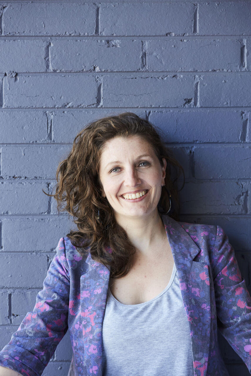 Belinda is standing against a blue painted brick wall. She is looking at the camera, smiling. She has long brown hair and is wearing a grey singlet top with a pink and purple blazer over top.