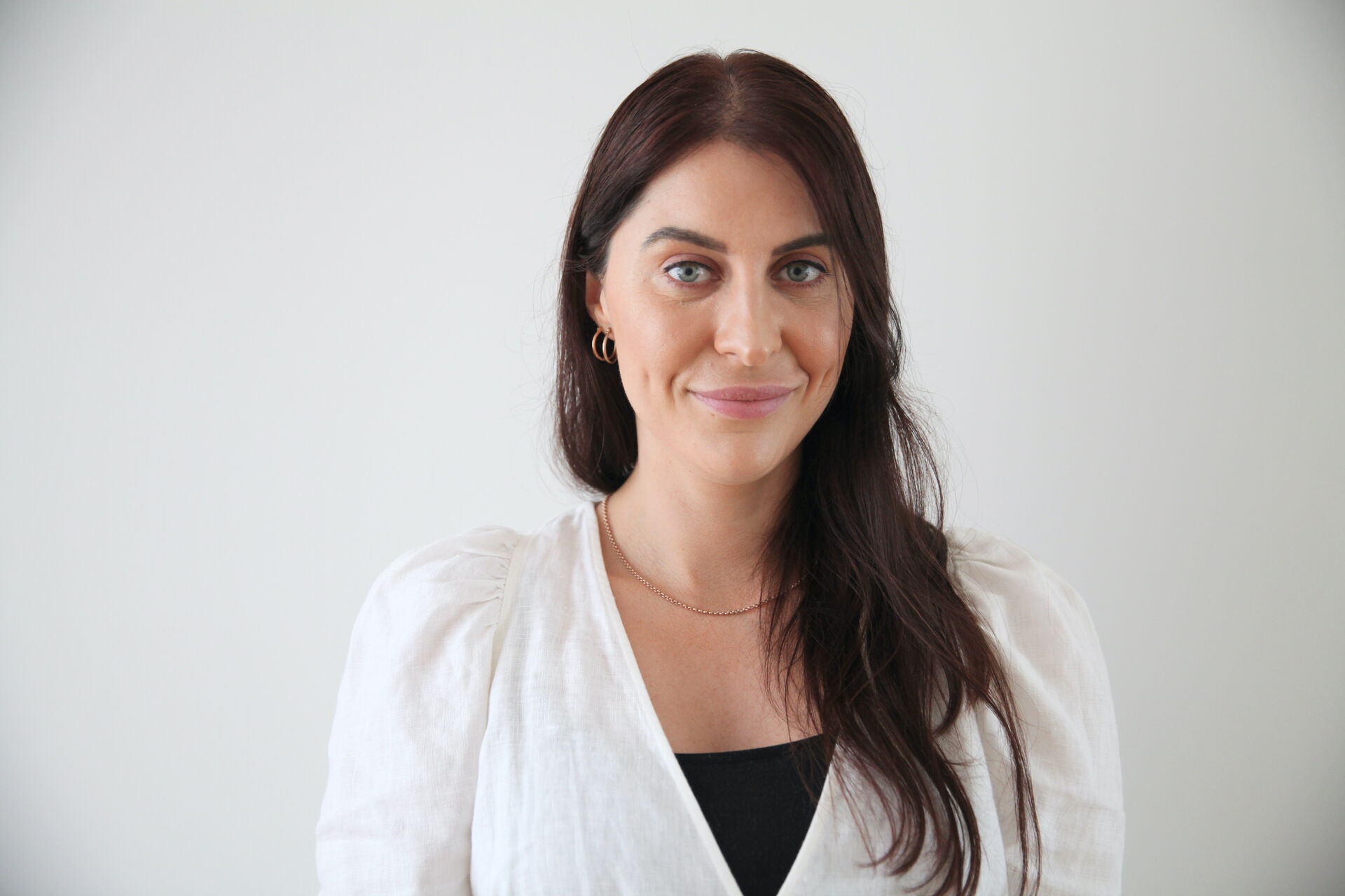Carly is looking at the camera, smiling. She has long dark hair and is wearing a white top with a black singlet top underneath.