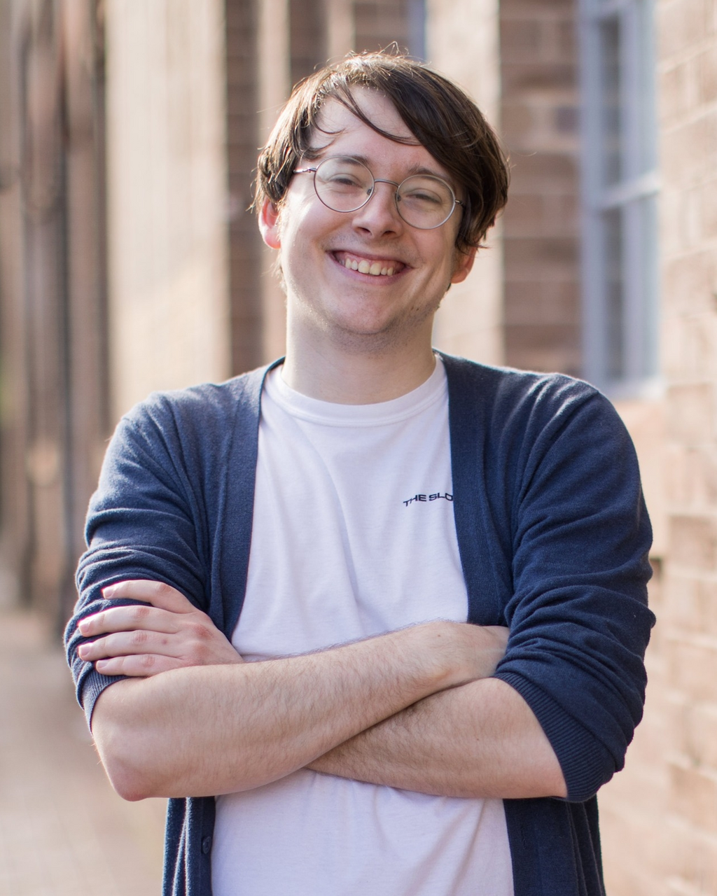 Ceri is standing outside with a building in the background. He is looking at the camera smiling, with his arms crossed. He is wearing a white tshirt, blue cardigan and glasses.