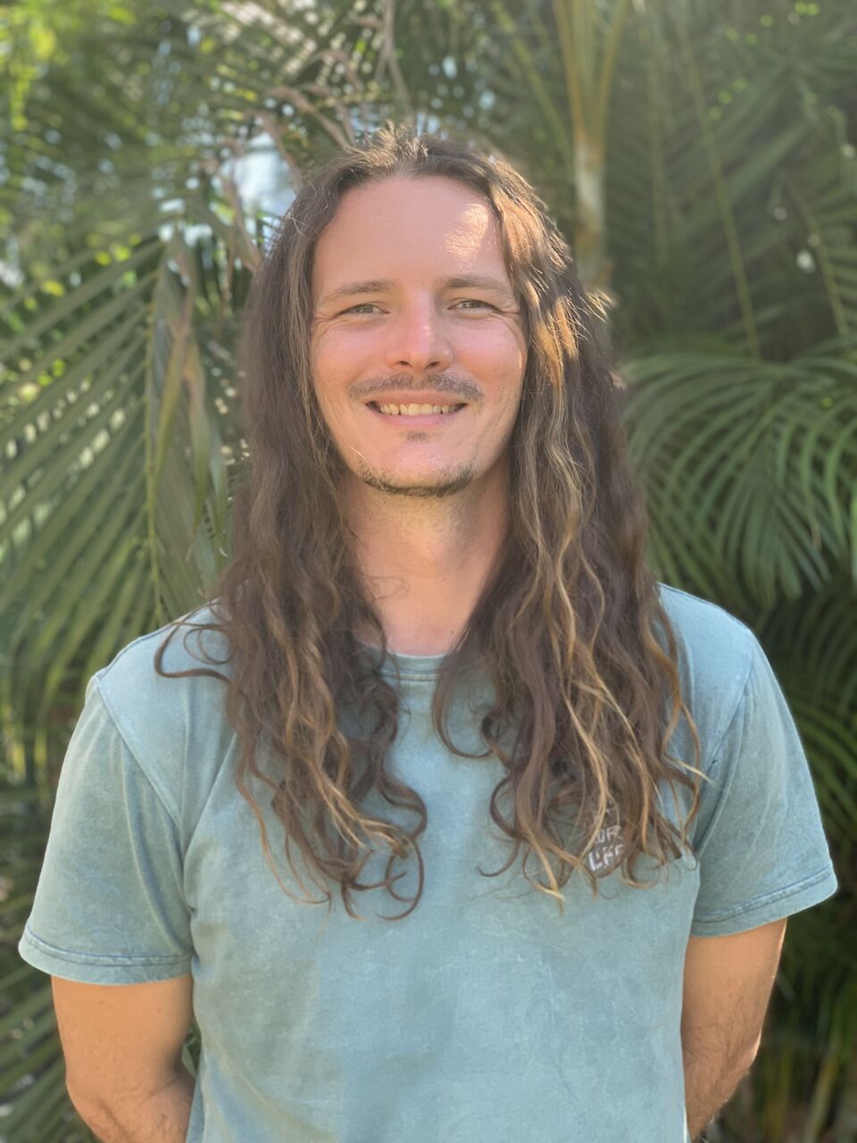 Dylan is standing outside with palm trees in the background. He is looking at the camera and smiling. He has long brown hair and is wearing a blue tshirt.