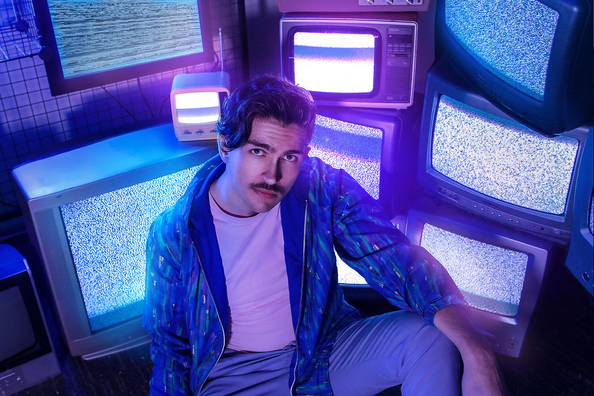 Jacob is sitting in front of some old television sets, stacked up, showing static screens. He has dark brown hair + moustache and is looking the camera.