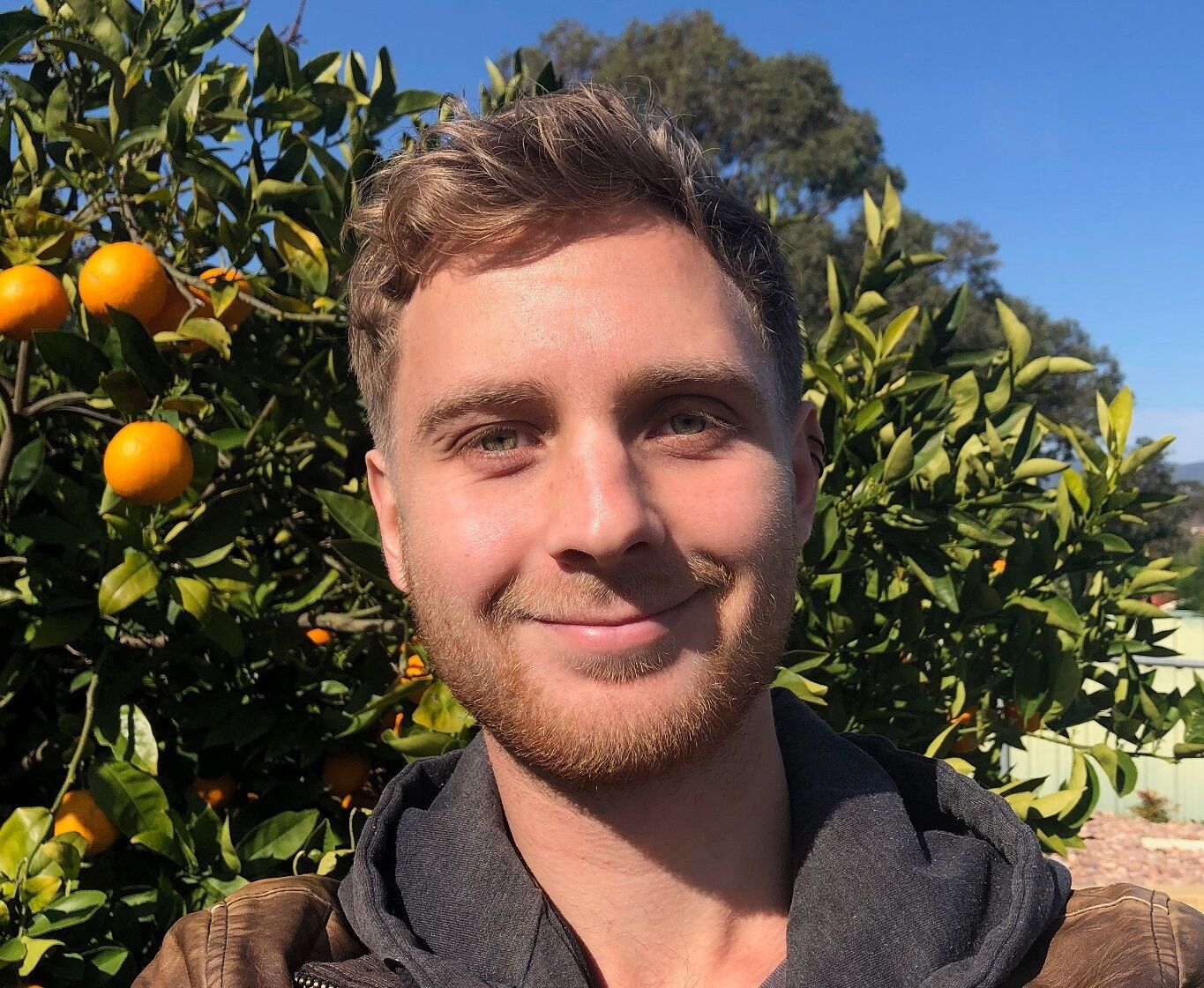 John is standing outside in front of a citrus tree. He has short light brown hair and is looking at the camera, smiling.