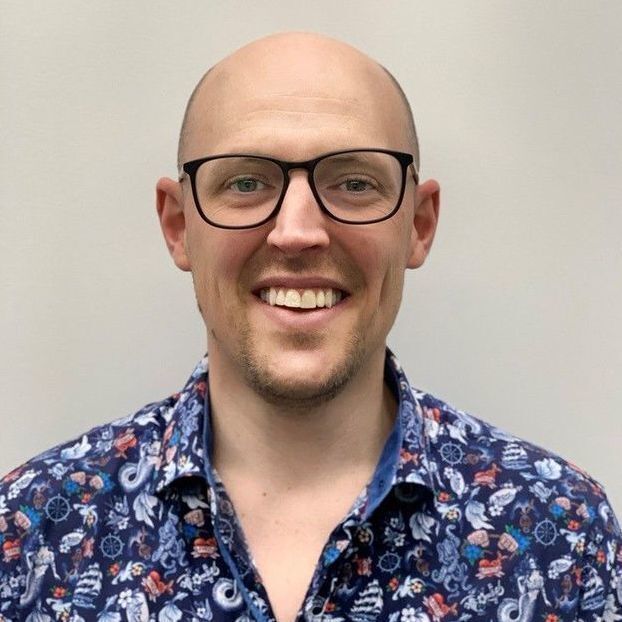 Luke Henry is looking at the camera, smiling. He is wearing a blue and red patterned button up shirt and black rimmed glasses.