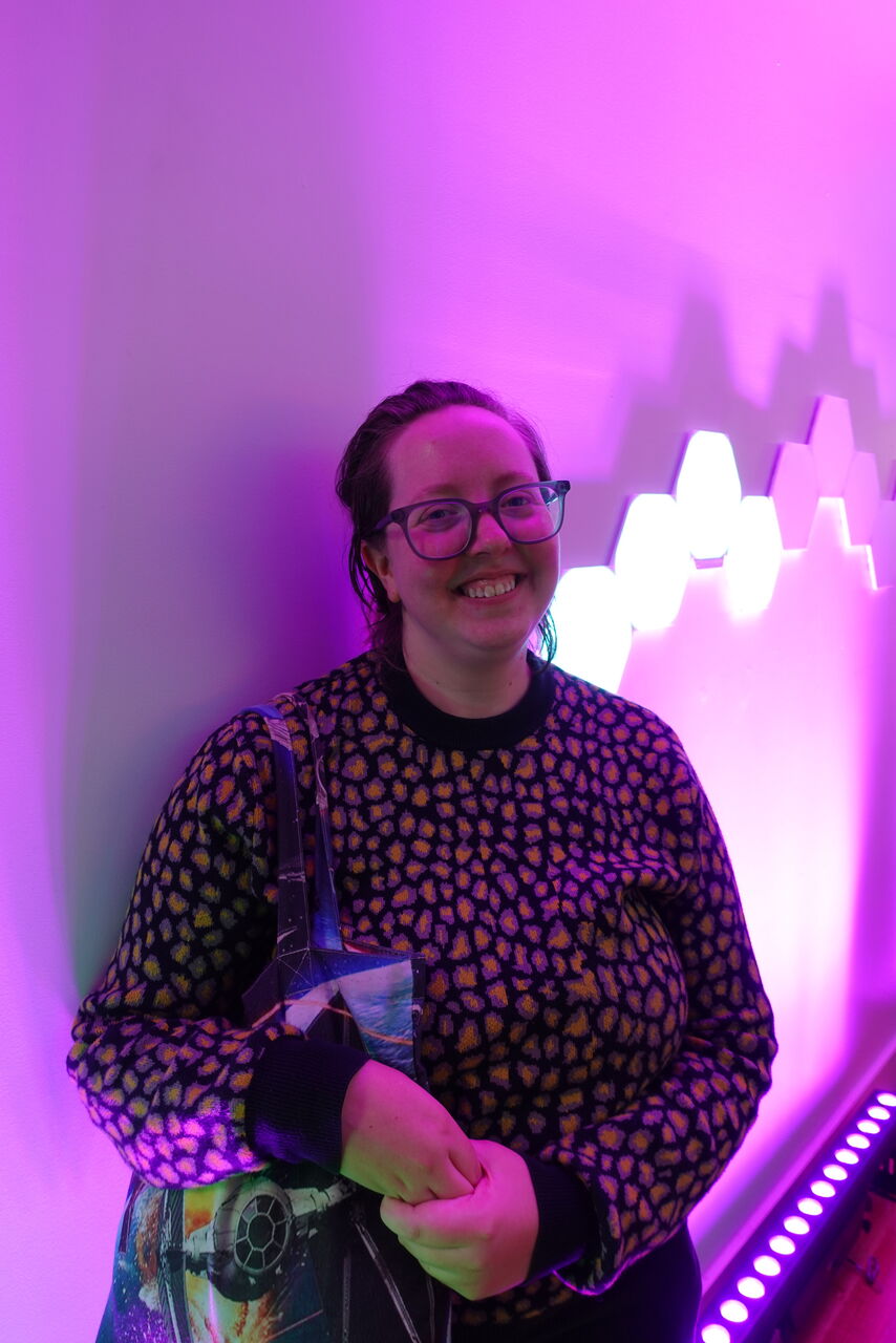Maize is standing in front of a purple fluro background. They are wearing a polka dot jumper, looking at the camera smiling.
