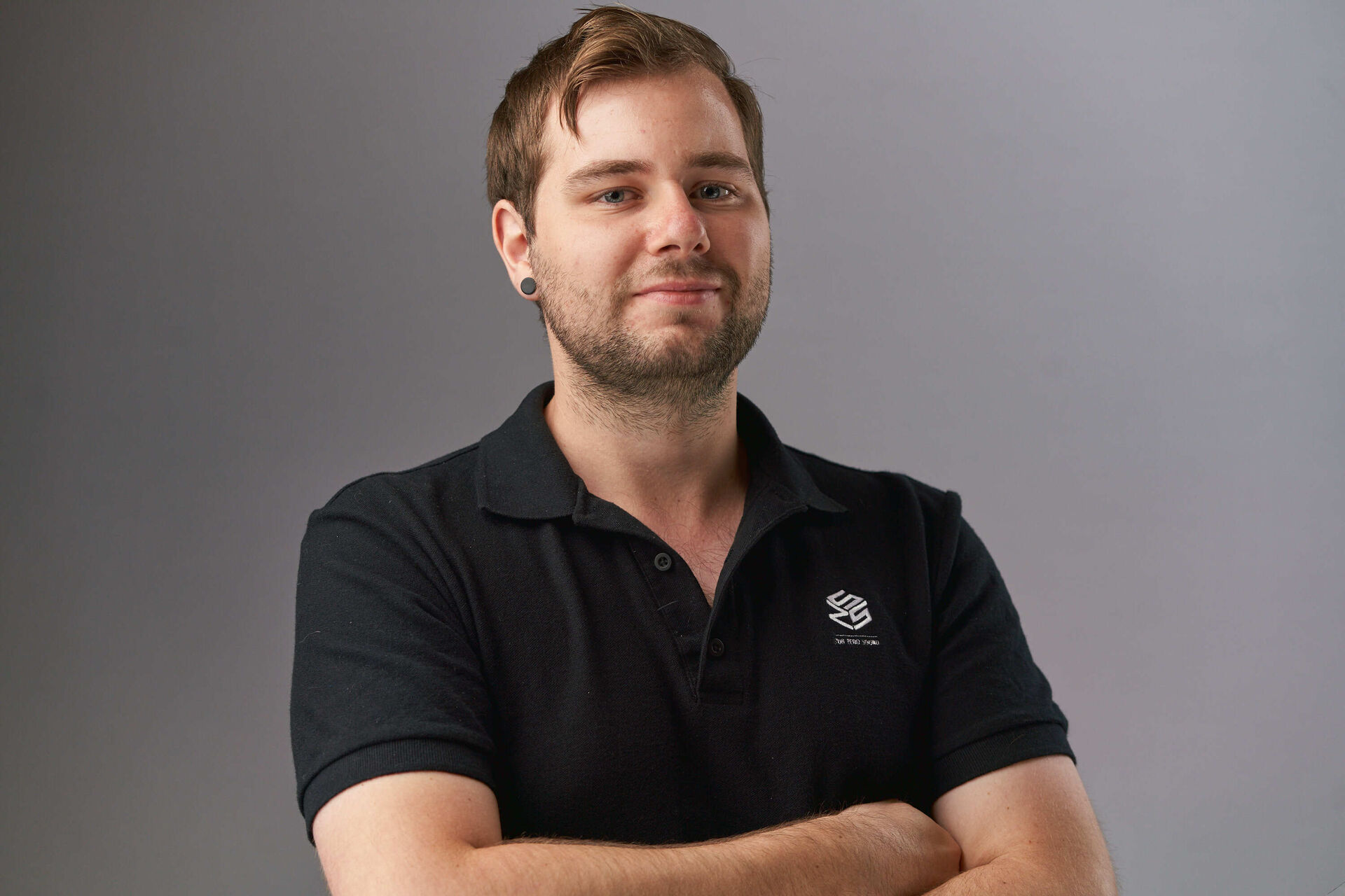 Matt is looking at the camera, with his arms crossed. He has short dark hair and is wear a black polo shirt.