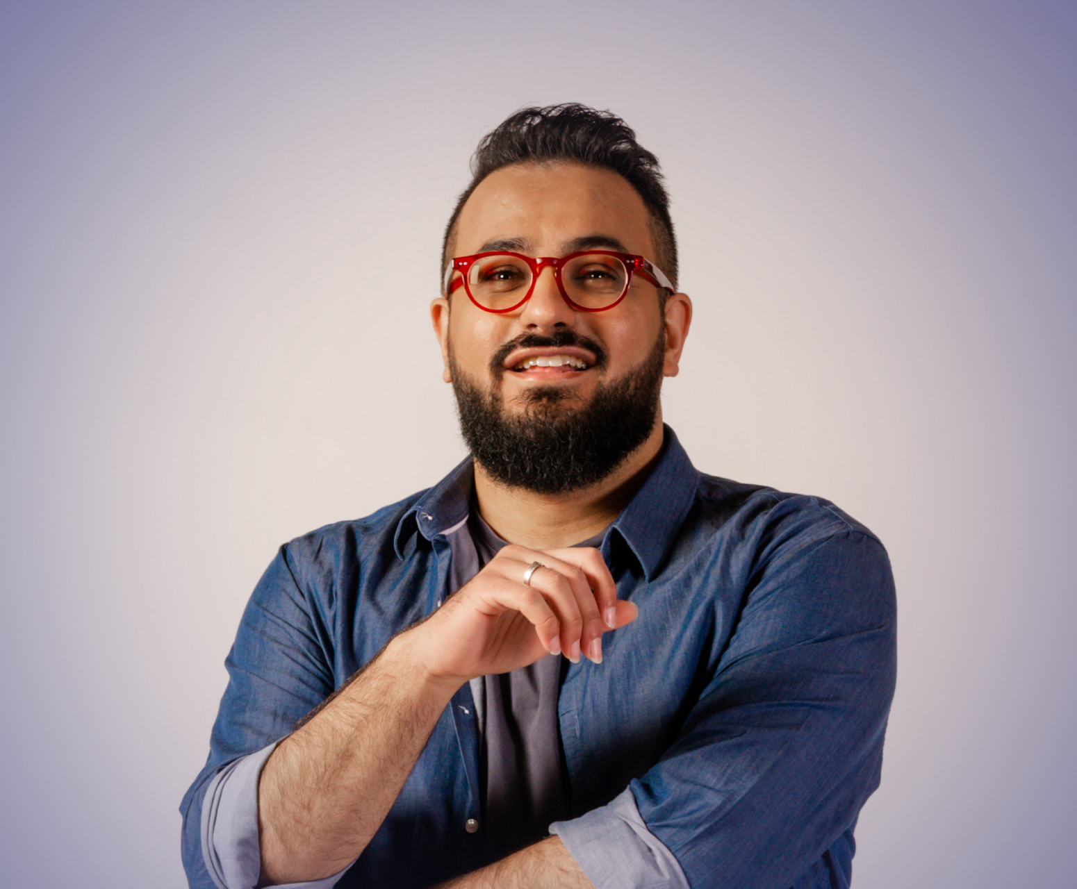 Meena is looking at the camera, smiling. He is wearing a button up denim top and red framed glasses.