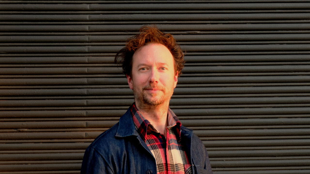 Michael is standing in front of a roller door, looking at the camera. He is wear a checked button up shirt and a denim overshirt.