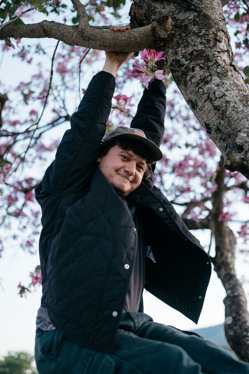 Narayana is outside, swinging from a tree. He is wearing a cap and dark coloured jacket. He is smiling at the camera.