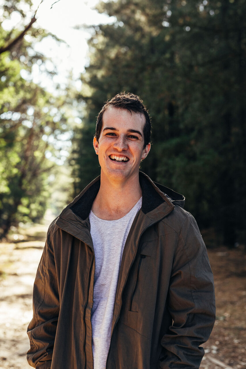 Scott is standing outside, looking at the camera smiling, with trees in the background. He is wearing a grey tshirt and a khaki jacket over the top.